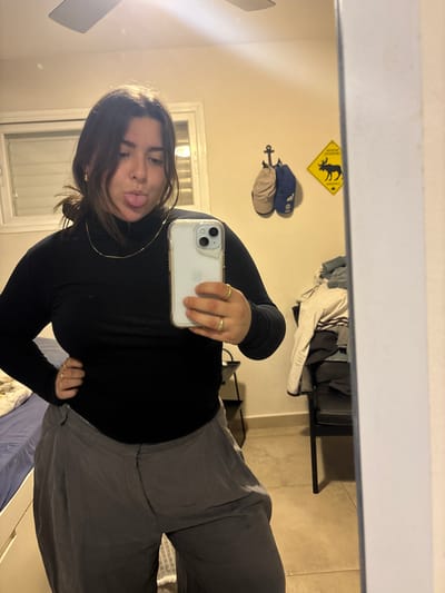 A person takes a mirror selfie, sticking their tongue out. They wear a black turtleneck and gray sweatpants. A gold necklace and rings are visible. The background includes a bedroom with beige walls, a window, a ceiling fan, and a yellow sign with a black moose. A coat rack with two hats and an anchor is also present. Clothes hang on a rack in the background. The image is taken with an iPhone.