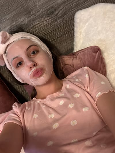 A young woman with a sheet mask and a pink headband with bunny ears is lying down on a wooden floor next to a plush white rug and a pink pillow. She is wearing a pink t-shirt with a daisy print. The scene appears to be indoors, possibly a bedroom or living room. There is no visible indication of the specific location in Bulgarovo, Bulgaria.