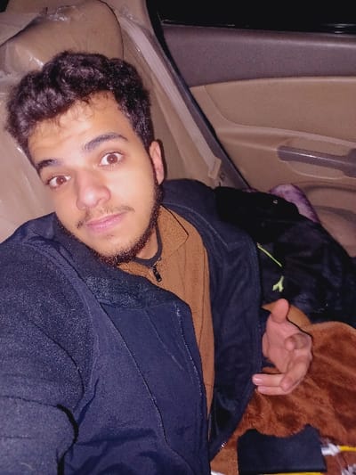A young man with curly dark hair, brown eyes, and a beard lies on a beige surface, seemingly inside a vehicle. He is wearing a brown sweater and a black jacket. Above him is a mix of items, including a black backpack with a green logo and a brown, fuzzy object. The vehicle's interior features a door panel with a handle. The overall lighting is dim.