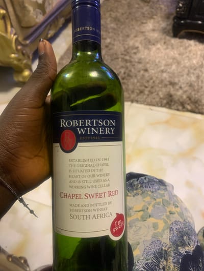 A close-up, slightly angled shot captured indoors in Akpajo, Nigeria, shows a dark-skinned hand holding a green glass wine bottle. The hand, adorned with a thin metallic chain bracelet on the wrist, firmly grips the bottle, revealing a dark mark or polish on the thumbnail. The wine bottle features a prominent label for "ROBERTSON WINERY," established in 1941, with a red "R W" logo. The label details "ESTABLISHED IN 1941 THE ORIGINAL CHAPEL IS SITUATED IN THE HEART OF OUR WINERY AND IS STILL USED AS A WORKING WINE CELLAR," and specifies "CHAPEL SWEET RED" wine, "MADE AND BOTTLED BY ROBERTSON WINERY SOUTH AFRICA," with a small red "i'm sweet" sticker at the bottom right. The background on the right reveals a blurred light-brown/beige shaggy carpet and a patterned fabric, possibly a garment, with blue, yellow, and white floral or abstract designs. The upper left background shows a golden, ornate, and blurred object, likely part of furniture or decor. The scene is illuminated by ambient indoor light.