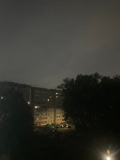 The image depicts a nighttime view of a residential building and surrounding area in Kirov, Russia. The building is a multi-story, grey apartment complex with numerous windows, many of which are illuminated, indicating occupancy. The sky is dark and overcast, suggesting cloudy weather. In the foreground, silhouetted trees obscure parts of the scene, with some foliage appearing wind-swept. Several cars are parked in the courtyard or on a nearby street. A bright light source, possibly a streetlamp, is visible in the lower right corner, emitting a strong glare. The overall impression is one of a typical urban residential environment at night, under subdued lighting conditions.
