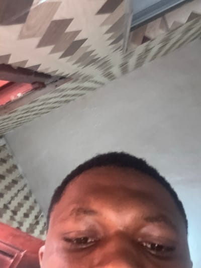 The image shows a close-up, angled view of a person's forehead and eyes, with part of a ceiling visible in the background. The ceiling has a pattern of light and dark brown geometric shapes. The person's skin tone is dark, and their eyebrows are visible. The lighting appears to be artificial and casts shadows on the ceiling. The image is low resolution and slightly blurry, making it difficult to discern finer details of the person's features or the environment. There is no identifiable information within the image to suggest a specific location in Obosi, Nigeria.