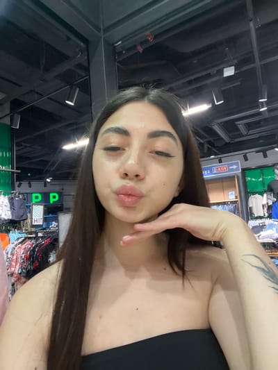 A young woman with dark hair and light skin, wearing a black strapless top, takes a selfie in a store. She is pouting and resting her chin on her hand, with a tattoo visible on her left arm. Behind her, racks of clothing are visible, along with store signage. The environment appears to be an indoor retail space. The signage includes the words "boys" and "A114", suggesting a section of a department store. The store lighting fixtures are visible on the ceiling. The overall scene indicates she is shopping.
