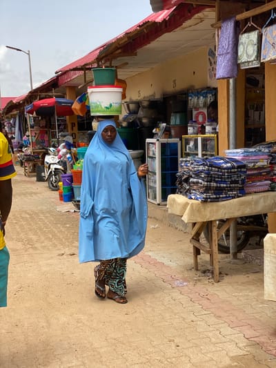A woman wearing a blue robe and head covering walks along a street in Kafanchan B, Nigeria. She is carrying several buckets stacked on her head. The street is lined with shops displaying merchandise, and a motorcycle is parked nearby. The ground is paved with reddish-brown and light gray rectangular bricks. The sky is overcast.