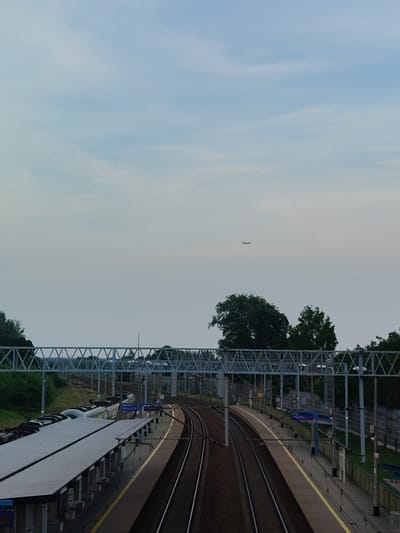 The image depicts a train station scene in Nowy Dwór Mazowiecki, Poland. The foreground features a covered train platform with a train parked alongside it, its sleek, modern design visible under the shelter. Multiple sets of train tracks curve away from the platform, disappearing into the distance. Overhead, a network of metal infrastructure, likely for power or signaling, forms a geometric pattern against the sky.
In the background, lush green trees punctuate the landscape, with one large, prominent tree to the right of center. Above the trees, a small airplane is visible in the pale, hazy sky, suggesting an airport might be nearby or that the plane is in transit. The overall lighting and the soft hues of the sky indicate that it is either early morning or late evening, possibly during an overcast day given the diffused light.
The scene appears to be devoid of people, giving it a sense of quietude. There is no overt activity taking place beyond the stationary train. The location is identifiable as a train station due to the platform, tracks, and overhead railway infrastructure. The weather appears calm, with a gentle, unobtrusive sky. No discernable text is visible in the image.