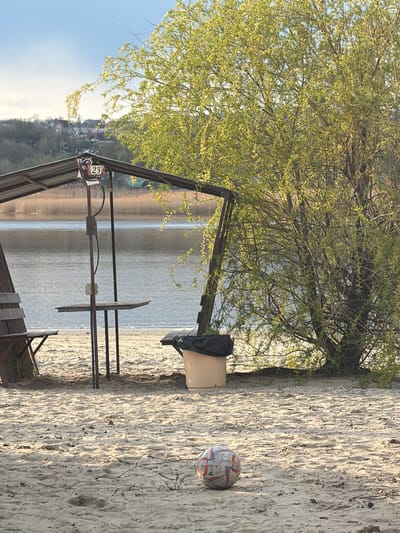 The image shows a sandy beach in Olginskaya, Russia, next to a body of water (likely the Don River, given the location). A metal structure with a sign labeled "23" stands near the edge of the beach. Behind it, there is a wooden box covered with a black plastic bag. A soccer ball rests on the sand to the right of the box. A tree with early spring foliage is located behind the box and the metal structure. The background shows a town/village on a hillside across the water. The weather appears sunny.