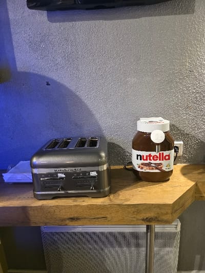 A large jar of Nutella is mounted on a wooden beam above a gray KitchenAid toaster. An electrical outlet is visible behind the Nutella jar. The beam also supports a wire-mesh structure with a metal bar. The wall behind the objects is gray with a textured surface. A dark object is visible on the left of the frame and a light blue light source illuminates the base of the wall.