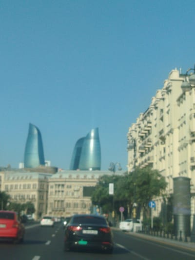 The image depicts a street scene in Baku, Azerbaijan, with a clear blue sky overhead. In the background, the Flame Towers, iconic skyscrapers that resemble flames, are visible. To the right of the Flame Towers, a long, multi-story building with many windows and balconies lines the street. In the foreground, cars are driving on a road. A red car is on the left, and a dark-colored car is in the right lane. Trees and streetlights are also present along the side of the road. The overall impression is that of a bright, sunny day in a modern city.
