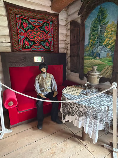 The image depicts a dimly lit interior scene, likely within a museum exhibit, showcasing a staged living room setting in what appears to be a traditional Russian izba (log house) in Ust'-Pyzas, Russia. A mannequin dressed in a light grey shirt and a patterned vest with black trousers is seated on a vibrant red couch. The mannequin has dark hair and a beard.
To the right of the couch, a round table is draped with a white lace tablecloth featuring intricate circular patterns and a decorative border. On the table, a silver samovar, a small glass with a silver holder, and a stack of papers with Cyrillic text are visible. A painting depicting a rural landscape with trees and small houses hangs on the wall behind the table.
Above the couch, a framed rug with a colorful floral and geometric design is mounted on the log wall. The walls themselves are constructed of rough-hewn logs. A white rope barrier, supported by simple wooden stands, separates the viewer from the exhibit. The floor is made of wooden planks. The overall impression is one of a preserved historical domestic interior.