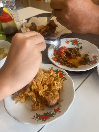 This is a close-up, overhead shot of a meal being shared at a table, likely in a restaurant in Medan, Indonesia. In the foreground, a person's left arm and hand are visible, holding a spoon. The spoon is hovering over a white plate filled with a golden-brown, crispy fried dish, possibly fried chicken or a type of fritter. The plate is decorated with small red and green floral patterns.
To the right of this plate is another white plate containing a vibrant red chili sauce or sambal, mixed with green chilies and oil. A metal spoon is partially submerged in this sauce. Further back, out of focus, is another dish of food, appearing to be fried meat, served on a piece of white paper. A clear glass of water and a condiment bottle with a red cap and yellow spout are also present on the table. The table surface is white, and the background is blurred, suggesting an indoor setting. The presence of sambal and the style of the fried food suggest it is Indonesian cuisine.