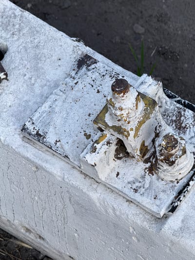 This is an extreme close-up, overhead shot of rusted metal components partially covered in white paint and some dirt. The main subjects are two bolts with nuts, one on the left and one slightly above and to the right. These bolts appear to be part of a larger, weathered metal assembly, possibly a bracket or hinge. The metal itself shows signs of significant wear, with rust visible in many areas, especially around the bolt heads and edges of the metal plates. The white paint is peeling and flaking, revealing the underlying yellow and brown metal.
The scene is outdoors, with a dark, granular surface, likely dirt or gravel, visible in the upper left and lower right corners of the image. A few blades of green grass are also peeking through the dark ground on the left. The lighting suggests it is daytime, but there are no clear indicators of specific time or weather conditions. The focus is entirely on the texture and condition of the metal hardware, highlighting its age and exposure to the elements. There is no text visible in the image. The overall impression is one of disuse and decay.