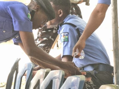 The image shows a scene in Maiduguri, Nigeria (latitude: 11.867, longitude: 13.123) with three men in blue uniform, possibly police officers. Two of them are bending down, with one pouring water from a plastic bottle onto a black object on the ground, while the third one observes them. They are situated in an outdoor setting, with a row of blue chairs in the foreground. The background is out of focus but appears to be a partially covered outdoor space, possibly a courtyard or a public area.