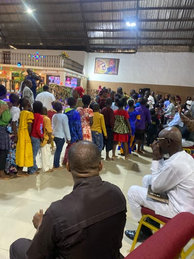The image depicts an indoor gathering of people, likely in a church or community hall, on Rukuba Road, Nigeria. A diverse group, including men, women, and children, are present, many dressed in brightly colored clothing. In the foreground, two men are seated, one in a black shirt facing away from the camera, and another in a white outfit with glasses, holding a book. Behind them, a crowd of people are standing, some with their backs to the viewer. Children are visible in the mid-ground, arranged in a line. The environment appears to be a large hall with a corrugated metal roof and exposed beams. A balcony with decorative lights and railings is visible in the background, along with a framed picture on the wall featuring a lion and two men. The lighting suggests artificial indoor illumination.