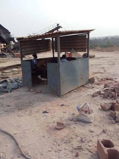 The image displays an outdoor industrial or construction site situated in the city of Hoche, Nigeria, under bright, possibly hazy daylight conditions. A makeshift, open-fronted shelter, constructed from corrugated metal panels for its lower half and horizontal wooden slats for its upper sides and roof, occupies the central mid-ground.

Inside the shelter, two adult males are seated. The individual on the left, positioned towards the front opening, wears a dark uniform with light blue shoulder patches, red stripes on dark trousers, a dark cap, and white athletic shoes, with his feet resting on a light-colored concrete block. The individual on the right is partially visible, wearing a light-colored shirt and seated further back within the structure.

The ground around the shelter and extending throughout the scene is unpaved, composed of dry, light brown soil, gravel, and scattered debris. This debris includes numerous fragments of red brick, broken concrete blocks, and a partially filled, light-colored fabric bag in the immediate foreground to the right of the shelter. A coiled dark hose or cable is visible on the ground in the far left foreground.

The background to the left features large-scale industrial machinery, including metal conveyor belts, structural frameworks, and access stairs, indicative of a material processing operation. Piles of rebar and other construction materials are scattered on the ground near these structures. The background to the right shows an extensive expanse of unpaved, disturbed ground with scattered dirt mounds and a large, dark cylindrical object lying horizontally. A dense line of green vegetation forms the distant horizon.