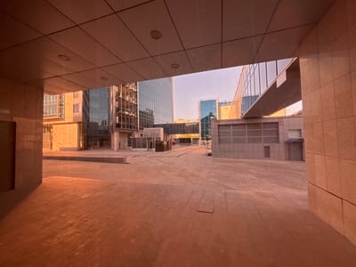 The media file shows an upside-down view of a modern urban setting, likely Orenburg, Russia. The foreground features a tiled walkway or platform, its geometric pattern illuminated by a warm, orange-tinted light, suggesting it's either sunset or sunrise, or artificial lighting. The tiles are large and square, with some circular indentations visible, possibly light fixtures.
Beyond the foreground, the scene opens up to reveal a complex of modern buildings with glass facades that reflect the sky and surrounding architecture. These buildings are tall and appear to be offices or residential complexes, characterized by their clean lines and contemporary design. One building in the mid-ground has a distinct pattern of vertical and horizontal window frames. Another, further back, displays a more intricate lattice-like structure on its exterior.
The sky visible between the buildings is a pale, washed-out color, indicating daylight, though the warm lighting on the foreground tiles creates a sense of either dusk or dawn. The overall atmosphere is one of urban modernity, with a focus on architectural forms and reflective surfaces. There are no people or vehicles visible, making it a serene, almost deserted urban landscape. The perspective, being upside down, adds an unusual and disorienting quality to the image. The architectural style and general urban density suggest a relatively developed area within a city.