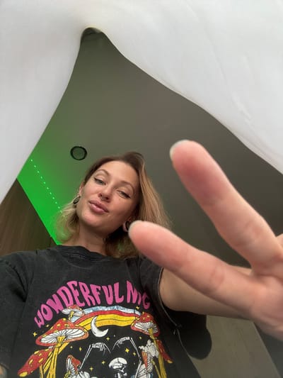 A woman is posing for a selfie from a low angle, her face is tilted upwards and her hand is extended towards the camera, making a peace sign. She has blonde hair, dark eyeliner, and a nose ring. She is wearing a black t-shirt with a colorful, psychedelic mushroom design. The background features a green LED strip light on a dark wall, and a white curtain or sheet is draped in the upper portion of the frame. The image appears to have been taken indoors, likely in a residential setting, and there is no indication of the location being Norilsk, Russia.