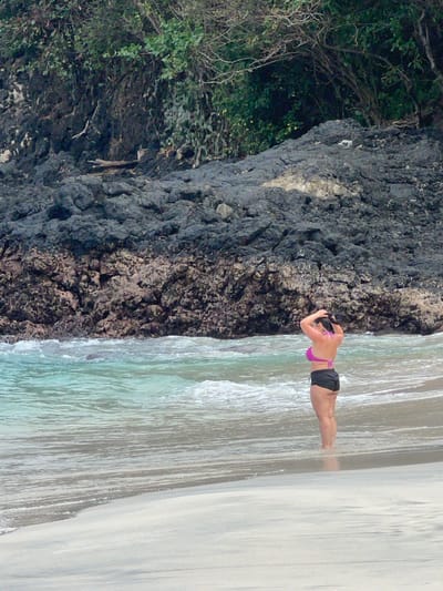 A woman in a purple bikini top and black shorts is doing a push-up on a sandy beach. Her body is parallel to the ground, and she is facing towards the water. The water is a clear, turquoise color and appears shallow. In the background, there is a rocky cliff face covered in lush green vegetation. The scene is set in a tropical location, likely during the day, given the bright lighting. The woman's posture suggests she is engaged in a physical activity, possibly a workout or a playful pose. There is no visible text in the image. The overall atmosphere is serene and natural. The location appears to be Manggis, Indonesia, known for its beautiful beaches and volcanic landscapes.
