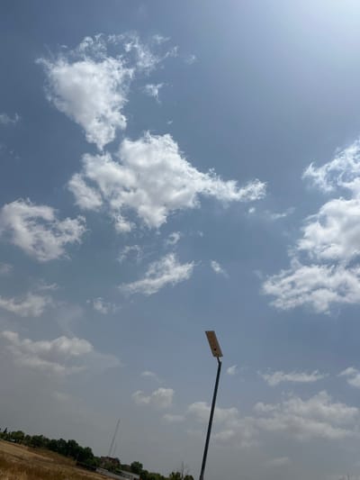 The image depicts an outdoor daytime scene in Baganakwo, Nigeria, under a bright, partially cloudy sky. The upper two-thirds of the frame are dominated by a clear blue sky interspersed with scattered white cumulus clouds of various shapes and sizes. A prominent subject is a tall, dark-colored pole, positioned slightly to the right of the center, extending from the bottom edge upwards. Mounted at the top of this pole is a rectangular, light-colored solar-powered street light fixture, angled slightly upwards, with visible solar panels on its upper surface. In the lower left quadrant, the foreground consists of dry, yellowish-brown ground with sparse vegetation. Further back, a horizontal line of green trees and bushes is visible, behind which indistinct structures and a tall, thin antenna or mast can be discerned in the mid-distance. No individuals are present in the frame.