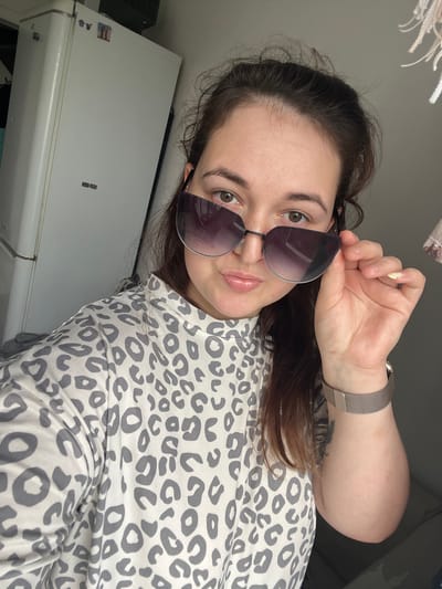 The image shows a young Caucasian woman with brown hair and green eyes, wearing large, dark-tinted sunglasses and a grey and white leopard-print shirt. She is holding the edge of her sunglasses with her right hand. A silver mesh watch is visible on her left wrist. To her left is a white refrigerator, possibly in a kitchen or utility area. The background is a plain, grey wall. There is no indication of the city of Chelyabinsk, Russia, in the image.