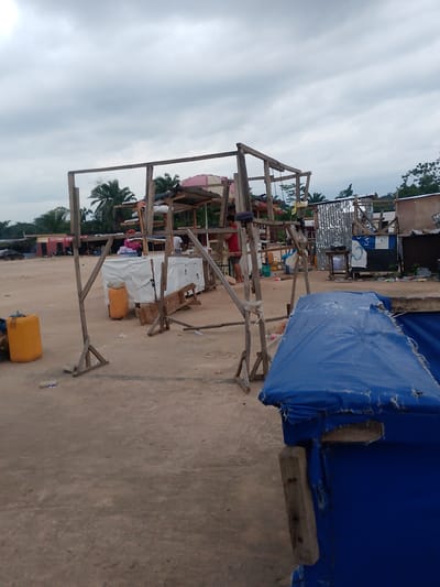 The image depicts an outdoor scene with market stalls in Egbagu, Nigeria. In the foreground, a damaged blue tarp covers a structure. A wooden beam is propped against it. To the left, a yellow plastic barrel is visible. In the middle ground, a wooden frame supports a structure covered with white sheeting, behind which a person in a red shirt stands. Further back, more market stalls constructed from wood and corrugated metal are visible, some with goods displayed on tables or shelves. A few people are present, tending to the stalls. The ground is unpaved, appearing to be dirt or sand, and scattered debris is present. In the background, trees and buildings can be seen under a cloudy sky. The overall impression is of a functional but somewhat makeshift market environment.