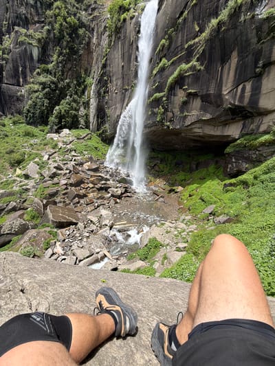 The image depicts a person's legs and feet resting on a large rock in the foreground. The individual is wearing black athletic shorts and a black knee brace on their left leg. They are also wearing hiking shoes with black and tan accents. The background features a powerful waterfall cascading down a rocky cliff face covered in lush green vegetation. The area at the base of the waterfall is filled with large, wet rocks and smaller stones. The environment appears to be a natural landscape, likely in the mountains. The location is identified as the Rohtang Waterfall in Bahang, India.