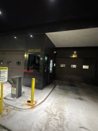 This image depicts the nighttime entrance of a private garage facility in Boston, United States. A specific street address or neighborhood within Boston cannot be determined from the visual information provided.
The scene focuses on a dark-colored building structure, likely a parking garage entry point, illuminated by artificial overhead lights. A central security booth, constructed with dark, reflective panels and featuring windows, is visible. Above the booth's window, a gold sign with dark lettering reads "PRIVATE GARAGE." To the immediate left of the booth is a grey metal parking arm mechanism or barrier post. Adjacent to this mechanism, a yellow-and-white "CAUTION" sign is prominently displayed, partially readable as "GATE ARM IN MOTION" and "DO NOT WALK AROUND VEHICLES!".
Several yellow cylindrical bollards are installed in the concrete ground in front of the parking arm and near the booth, with some displaying reddish-brown rust stains at their bases. A dark, possibly wet, hose or conduit runs along the ground from the area of the parking arm towards the garage entrance. To the right of the security booth, a large, dark grey roll-up garage door is visible, featuring multiple small, rectangular windows across its width. Above the garage door, a white rectangular sign with yellow lettering is affixed to the wall. At least two security cameras are mounted on the building's facade, one near the security booth and another further right, above the garage door. The concrete ground surface shows signs of wear and appears to have damp patches or stains. No individuals, vehicles, or active interactions are present in the image.