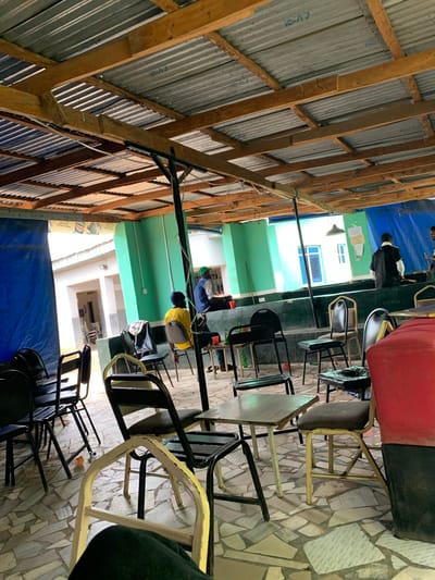 The image depicts an outdoor covered area with a corrugated metal roof supported by wooden beams and metal poles. The floor is tiled with irregular stones. In the background, there is a green building with white trim and windows. A blue tarp hangs on the left side, and a red object, possibly a large cushion or seating, is visible on the right.
Several people are present. On the left, a person wearing a yellow shirt and a dark beanie sits facing away from the camera, while another person in a blue vest and green hat is seated further back on a bench. In the middle-right, two individuals are near a counter. One, wearing a black uniform, is seated, and another, dressed in dark clothing, stands nearby.
Scattered throughout the space are various chairs, mostly black metal or with dark seats and frames, and some with light-colored frames. Small tables are also present, some with drinks or objects on them. A plastic bottle is visible on the floor in the foreground. The overall impression is that of a casual, perhaps open-air, seating area or eatery in New Stadium, Nigeria.