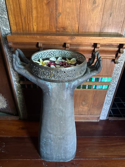 A metal sculpture of two cupped hands holds a decorative bowl filled with plumeria flowers. The sculpture is positioned in front of a wooden wall panel, with intricate silver trim visible on either side. Below the bowl, a stained-glass panel features horizontal bands of green and blue glass. The sculpture and bowl appear to be made of aged metal with a patina. The scene is set indoors, likely in a hotel or resort lobby in Baan Lamai, Thailand, given the typical decorative elements found in such establishments in the region.