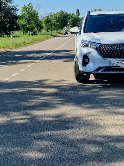 In Kursava, Russia, a white Haval SUV is parked on the shoulder of a two-lane asphalt road under a clear blue sky. The front of the vehicle is prominently featured, with its grille and headlight visible. The license plate reads "A 727". To the left of the road, lush green grass and trees line the landscape. In the distance, a person is riding a purple motorized scooter on the opposite side of the road, heading away from the camera. The sun casts dappled shadows of tree branches across the road and the front of the SUV.