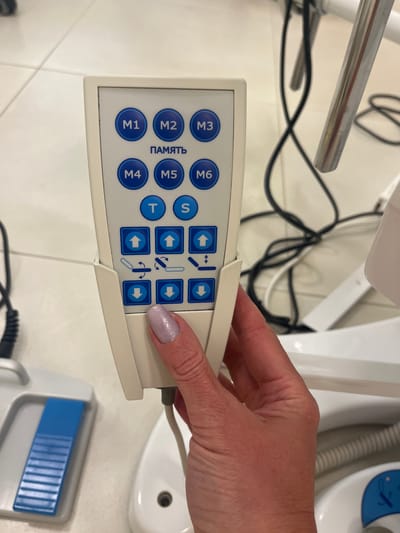 In an indoor environment with tiled flooring, a person's hand with light purple nail polish is holding a beige medical device control panel. The control panel features multiple blue circular buttons labeled "M1" through "M6," a button with the Russian word "ПАМЯТЬ" (memory), and buttons labeled "T" and "S". Below these, there are square buttons with directional arrows: up, down, and two symbols indicating adjustment or rotation. A blue foot pedal is visible in the lower left corner of the frame. The background includes white medical equipment and black cables. The exact location in Kaliningrad, Russia, cannot be determined from the provided image.