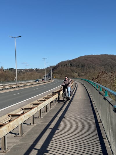 The image depicts a daytime scene on the Ruhrtalbrücke (Ruhr Valley Bridge), which carries the A43 autobahn in Hattingen, Germany. The sky is clear and bright blue.

In the foreground and midground, a multi-lane highway, paved with asphalt, curves from left to right into the distance. It is bordered on its right by a rusted, multi-beam metal guardrail system. To the right of this guardrail is a concrete pedestrian and bicycle path.

A single adult female is present on this path, positioned towards the right side of the frame. She is Caucasian, wearing dark sunglasses, a dark brown or grey jacket, blue jeans, and light-colored sneakers. She is walking forward and pushing a dark-framed bicycle with both hands on the handlebars. The bicycle is equipped with a front bag or basket and two dark panniers on a rear rack.

The pedestrian path is bordered on its outer right edge by a lighter metal railing with a green top bar. Long, distinct parallel shadows from the guardrails and railings are cast across the path, indicating bright sunlight from the left.

Multiple streetlights with double-arm fixtures are spaced along the highway. Several vehicles, including cars, are visible on the highway in the distance, moving away from the camera's perspective.

The background consists of rolling hills. The hill on the right is densely covered with bare deciduous trees, suggesting late autumn, winter, or early spring. The hill on the left features scattered trees and some residential structures.