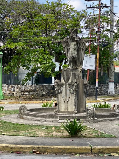 The image presents an outdoor urban scene in Valencia, Venezuela, centered around a prominent, weathered stone monument. The monument features a standing, sculpted human figure, possibly a religious or historical personage, with a lean physique and flowing drapery. This figure rests atop an ornate base, which is adorned with detailed carvings resembling winged heads or grotesque faces, and decorative scrolls.
To the right of the monument, a paved area, likely a sidewalk, extends alongside a narrow strip of grass. A yellow-painted curb marks the boundary with what appears to be a street or road. Small green plants are scattered in the grassy sections and around the base of the monument.
The background is filled with large, leafy green trees, casting shade over parts of the scene. Above and intertwined with the tree branches, a dense network of black electrical or communication wires crisscrosses the frame, supported by several utility poles. One pole is noticeably orange-red and bears yellow lettering, with "UCEQ" and "266" clearly visible. A white rectangular sign is attached to a pole adjacent to the monument, displaying the Spanish word "AQUI" prominently at the top of its text block, followed by smaller lines of text. Further back, a stone wall and metal fences are visible, suggesting nearby buildings.
The scene is captured during daytime, under an overcast sky. No discernible human activity is taking place. The monument's aged appearance and the intricate overhead wiring are notable details of this public space.
