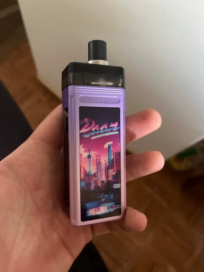 A person's hand holds a purple and silver vape device. The vape device has a black mouthpiece on the left side. The main body of the device is a light purple with a decorative sticker on the front. The sticker features a stylized cityscape with tall buildings and a vibrant pink and blue sunset. The word "Dubai" is written in large, stylized, pink lettering across the cityscape. The background shows a white wall and a wooden floor. The lighting suggests it is indoors during the day. The image appears to be a close-up shot, focusing on the vape device and the hand holding it.