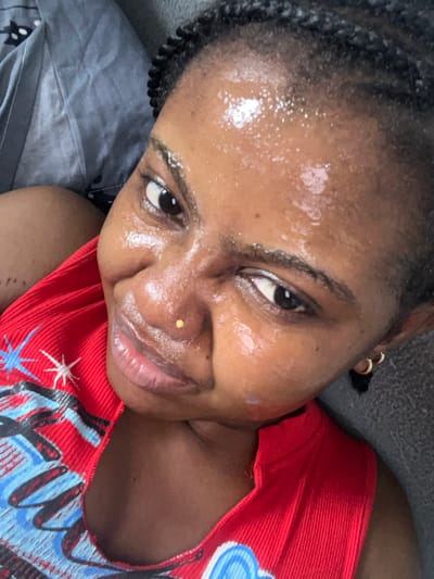 A female individual is depicted in a close-up, high-angle shot within the city of Ipetumodu, Nigeria. Her dark skin is noticeably moist or oily across her forehead, nose, and cheeks, reflecting ambient light. Her dark hair is styled in multiple small braids. A small, gold-colored stud pierces her left nostril. Small, gold-colored hoop earrings are visible on her right earlobe. She wears a red, ribbed sleeveless top with a blue and white patterned design on the left side of the chest. Her eyes are dark and focused directly forward, with a slight, closed-mouth smile on her lips. A faint, light-colored mark is present on her left cheekbone. The background consists of a crumpled grey fabric, possibly bedding, to the left, and a darker, textured grey surface to the right.