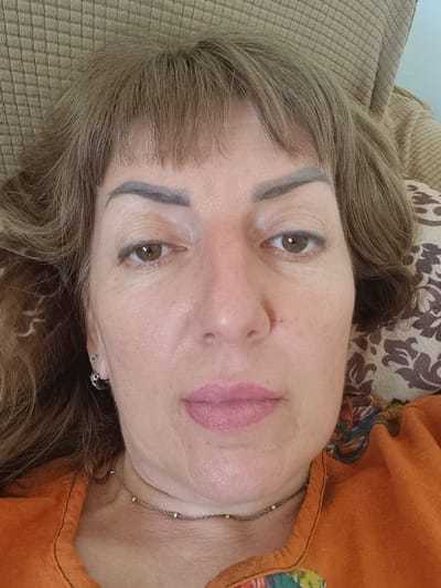A selfie shows a Caucasian woman in her late 40s or 50s with medium-length brown hair and bangs. She is lying down, with her head resting on a brown textured surface, possibly a sofa or chair. The woman is wearing an orange shirt with a patterned neckline and a delicate silver necklace. Her left ear is adorned with a silver hoop earring. She appears to be indoors, with a patterned pillow visible behind her head. The overall impression is a casual, unposed moment. The location is not specified in the image itself.