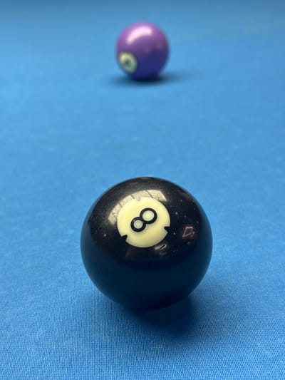 The image shows two billiard balls on a blue felt surface. The 8-ball is in the foreground, slightly to the right. A purple ball is in the background to the left, out of focus. The scene's location is unknown; there is no visual information to associate it with the city of Bellville, South Africa.