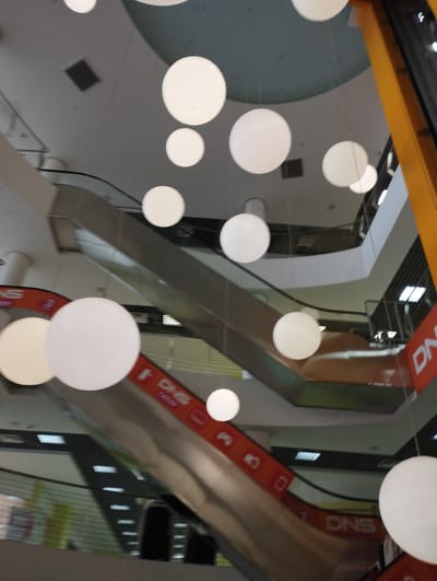 The image depicts an interior view of a multi-level shopping mall. Prominently featured are several spherical, white pendant lights hanging at various heights. These lights are the primary focus, illuminating the space with a soft glow. Below the lights, escalators are visible, suggesting the mall's vertical circulation. One escalator is seen in the foreground, with an orange banner featuring the text "DNS" and various icons. Other architectural elements of the mall, such as railings, ceilings, and walls, are also present, though some are out of focus or partially obscured by the lights. The overall impression is a brightly lit, modern commercial environment. The image is located in the city of Korolyov, Russia.