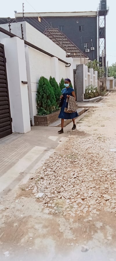 An image taken in Kaduna, Nigeria, depicts a female subject walking on an unpaved, uneven dirt path with scattered light-colored gravel and debris, moving from right to left across the frame. She wears a dark blue, short-sleeved dress, dark shoes, a blue head covering, and a light blue surgical mask. She carries a dark handbag on her left shoulder and a patterned fabric bag or item draped over her right forearm.
To the left of the subject, a tall, white perimeter wall with an electric fence system on top runs parallel to the path. A dark brown gate is partially visible on the far left. A security camera is mounted on this wall. Below the wall, three conical green evergreen shrubs are planted in a brick planter box. Further along, another section of a white wall with decorative architectural elements and an electric fence is visible.
In the background, a multi-story, dark grey or black building with multiple windows and mounted air conditioning units stands prominently. A lower building with a brown-tiled roof is situated in front of it. A tall, dark metal structure supporting a black water tank is visible on the far right. The sky is bright but appears hazy or overcast. Shadows from an unseen structure are cast on the ground to the left of the subject.