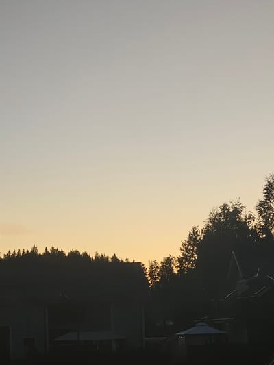 The image depicts a twilight scene with a silhouette of a forest and buildings against a pale yellow and grey sky. The time of day suggests either dawn or dusk, with the sun low on the horizon, casting a warm glow. In the foreground, dark silhouettes of what appear to be houses or outbuildings are visible, with some having pitched roofs. A small gazebo-like structure with a peaked roof is also present. Behind these structures, a dense line of trees forms a dark, undulating silhouette. The sky is largely featureless, with a gentle gradient of color from a pale yellow near the horizon to a light grey at the top of the frame. The overall impression is one of rural or suburban tranquility, with the natural landscape dominating the scene. The location is identified as Mäntsälä, Finland, which is a municipality in the Uusimaa region of southern Finland, known for its rural characteristics and proximity to natural landscapes.