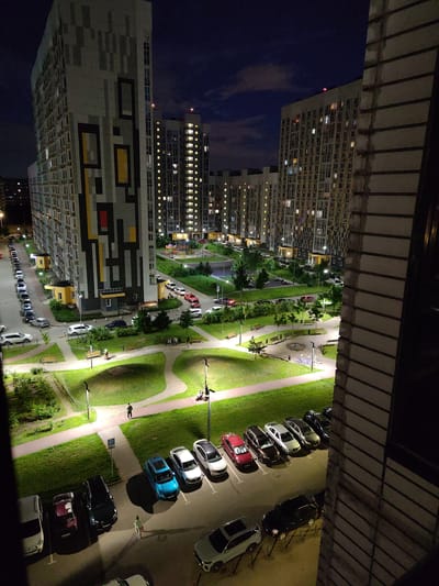This is a vertical, medium-range night-time photograph taken from a high vantage point, likely a balcony, overlooking a residential courtyard in Lyubertsy, Russia. The scene depicts a complex of modern, multi-story apartment buildings, their windows illuminated, suggesting that it is evening or night. The buildings feature diverse architectural styles, with one prominent building showcasing a striking geometric facade in shades of grey, black, yellow, and red.
Below the buildings, a well-maintained courtyard is visible, characterized by green lawns, paved pathways, and scattered trees and benches. Several cars are parked in designated areas, lining the roads and in a parking lot. The lighting in the courtyard, primarily from streetlights, casts a warm glow on the grass and pathways, creating areas of light and shadow.
There are a few people visible in the scene, appearing as small figures walking or standing in the courtyard. One person is walking along a paved path, while a small group is gathered near a bench. Further in the distance, near the buildings, there appears to be a playground with brightly lit equipment.
The sky above is dark blue, with hints of clouds, indicating it is night. The overall atmosphere is calm and serene, portraying a typical urban residential environment after dark. The image captures the transition from day to night in a modern urban setting.