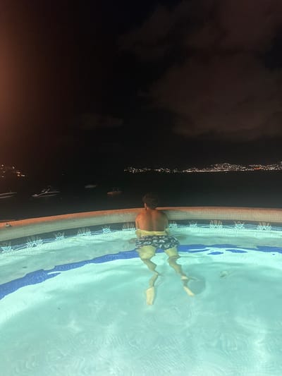 The image shows a swimming pool in Acapulco, Mexico, at night. A person wearing patterned swim trunks is in the pool, submerged from the waist down. The pool's edge is tiled with a floral pattern. The surrounding environment is dark, with some light reflecting off the water's surface.
