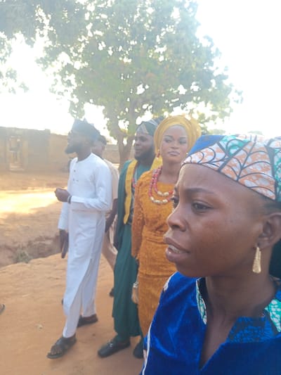 A group of people stand outdoors. One person wears a blue patterned headscarf and a blue top. A woman behind her has on a yellow head covering and a dress of the same color. A man stands next to her in a green top. Further to the left is another man in a white outfit and sandals. A tree with many branches fills the background, partially obscuring a building.