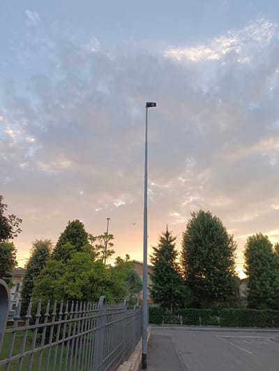 A street lamp stands tall in the center of the frame, its dark head silhouetted against a cloudy sky tinged with the warm hues of sunset. The sky transitions from a pale blue at the top to shades of orange and pink near the horizon. Wispy clouds are scattered across the sky, some catching the last light of the day.

In the foreground, a dark metal fence runs horizontally across the image, its pointed pickets creating a repeating pattern. Beyond the fence, lush green trees of various shapes and sizes create a dense backdrop. Some trees are tall and conical, resembling evergreens, while others are rounder and fuller. The foliage is vibrant and healthy, suggesting a temperate climate.

To the left of the fence, a patch of well-maintained green grass is visible. Further back, glimpses of houses with white walls and brown tiled roofs can be seen peeking through the trees, indicating a residential or semi-urban setting.

A paved road is visible to the right of the fence, with white lines marking a crosswalk. A faint "STOP" sign is visible on the road. The overall scene evokes a peaceful, tranquil evening in Viadana, Italy. The light suggests it is either early morning or late evening. The sky is cloudy, but the sun is still present enough to cast a warm glow. There are no people visible in the image.