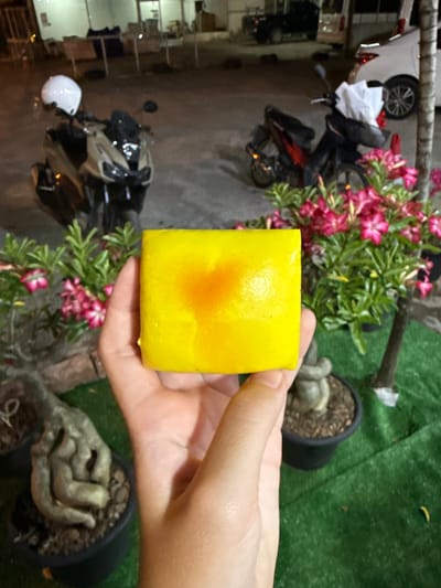 A hand holds a square-shaped yellow object, possibly a piece of food or a sponge, in front of a blurred background. Behind the hand, a bush with pink flowers is visible. To the left, a white car and a motorcycle are parked on a paved surface. The scene appears to be outdoors, possibly in a residential area or a shop's exterior, given the presence of vehicles and plants. The lighting suggests it might be late afternoon or evening, or an overcast day. No text is visible in the image. The location is identified as Baan Bo Phut, Thailand.