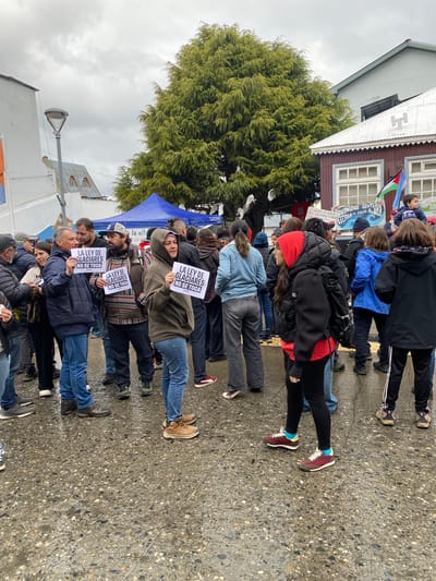 The image captures a protest in Ushuaia, Argentina. A group of people stand close together, holding signs that read "LA LEY DE GLACIARES NO SE TOCA" (The Glacier Law is Not Touched). They are standing on a wet, speckled pavement near a tree. Buildings are visible in the background. The sky is overcast, suggesting a cloudy day.