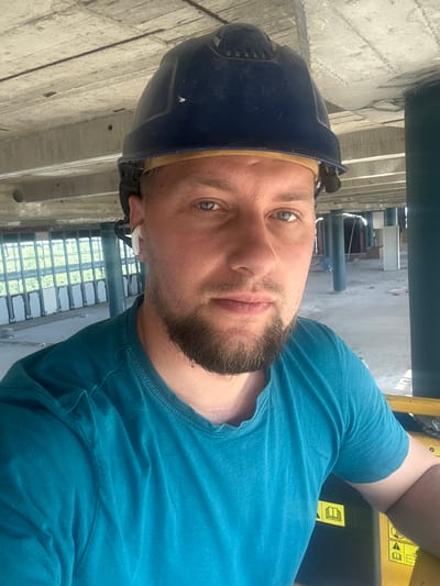 A mid-shot selfie shows a man wearing a blue hard hat and a teal t-shirt, standing in an unfinished building. He has short brown hair, blue eyes, and a short beard. He is wearing white wireless earbuds. The background shows concrete structures, including pillars and a ceiling with visible rebar. In the distance, there are windows with white frames and what appear to be industrial elements. The man is positioned in front of yellow machinery with warning symbols, suggesting he is on a construction site or a similar industrial setting. The lighting is bright, indicating daytime. The image does not contain any specific visual cues to pinpoint the location within Hamburg, Germany.