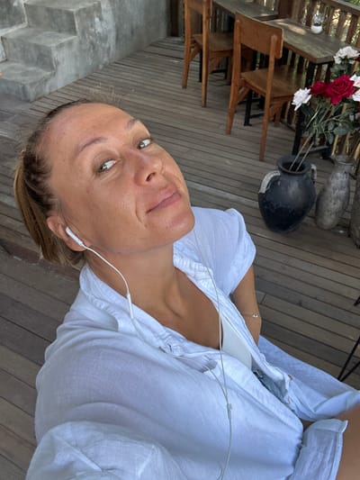 A selfie of a woman wearing a white shirt and white earphones. She is sitting on a wooden deck, with a concrete staircase and railing to her left. In the background, there are wooden tables and chairs, as well as a vase of flowers. The scene appears to be an outdoor cafe or restaurant. The location is not specified in the city of Kediri, Indonesia.
