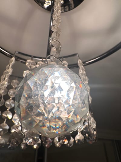 This is a close-up, eye-level shot of a clear, faceted glass orb, likely a decorative element of a chandelier or light fixture. The orb is round and appears to be made of many small, flat surfaces that catch and refract light, creating rainbow-like glints across its surface. It is suspended from a thin, metallic loop or ring.
Strands of smaller, clear glass beads, also faceted, are attached to the orb and trail away from it, connected by thin metal rings. These strands are arranged in a cascading fashion, suggesting they are part of a larger decorative piece. The background is out of focus and consists of muted, neutral tones, with a soft light source illuminating the main subject. The lighting suggests an indoor setting, possibly a room with ambient light or a light fixture that is on. There is no discernible text, people, or weather conditions visible. The overall impression is one of sparkle and delicate craftsmanship.