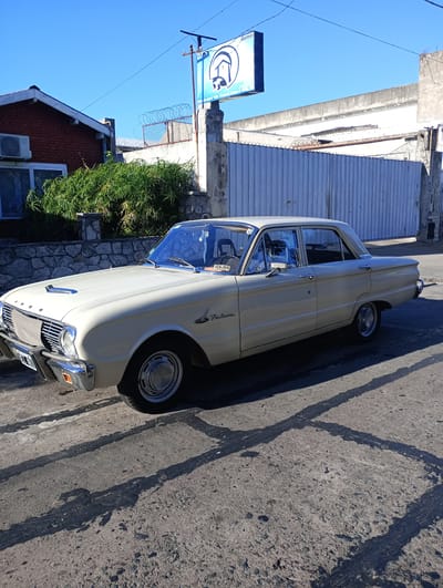 A vintage Ford Falcon automobile is parked on a weathered concrete street in Wilde, Argentina. The car is light beige, possibly cream, with visible chrome trim and a license plate. The background features a brick building with a window air conditioning unit, a stone wall topped with greenery, and a high, corrugated metal fence. A blue sign with white lettering and a graphic stands near the fence. The lighting indicates a sunny day, casting stark shadows from the car and surrounding structures onto the cracked pavement.