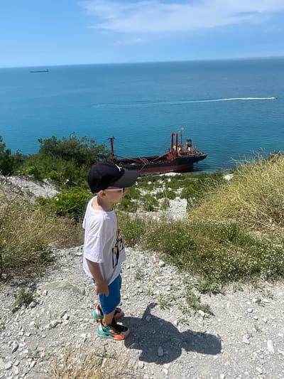 The image depicts a young boy wearing a white t-shirt, blue shorts, a black baseball cap, and colorful sneakers, standing on a rocky, vegetated incline overlooking the sea. In the distance, a grounded cargo ship, appearing rusty and partially submerged, rests near the shore. The sea is a vibrant turquoise, with a boat leaving a white wake in the middle ground and a distant ship on the horizon. The sky is blue with scattered clouds. The scene is likely located near Novorossiysk, Russia, a city known for its coastline and maritime activity. The overall impression is one of a sunny day by the sea, with a unique, shipwrecked element adding to the landscape.